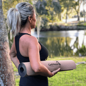 A blonde woman stands in a warm weathered marsh wearing athletic clothes and carrying a rolled cork yoga mat. 