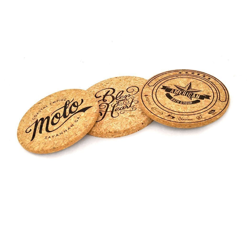 Three custom printed coasters with black printing. The furthest left coaster says "moto coaster empire, savannah ga". The middle coaster says "bless your heart" in a flowing cursive script. The coaster on the right says "American Bath Group" with a star in the center.