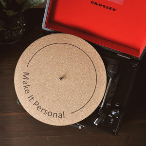 A natural cork turntable mat with a black circular line with "make it personal" written on the mat. This mat is on a black suitcase turntable mat with a red interior.