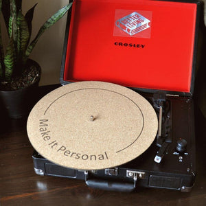 A natural cork turntable mat with a black circular line with "make it personal" written on the mat. This mat is on a black suitcase turntable mat with a red interior on a brown table.
