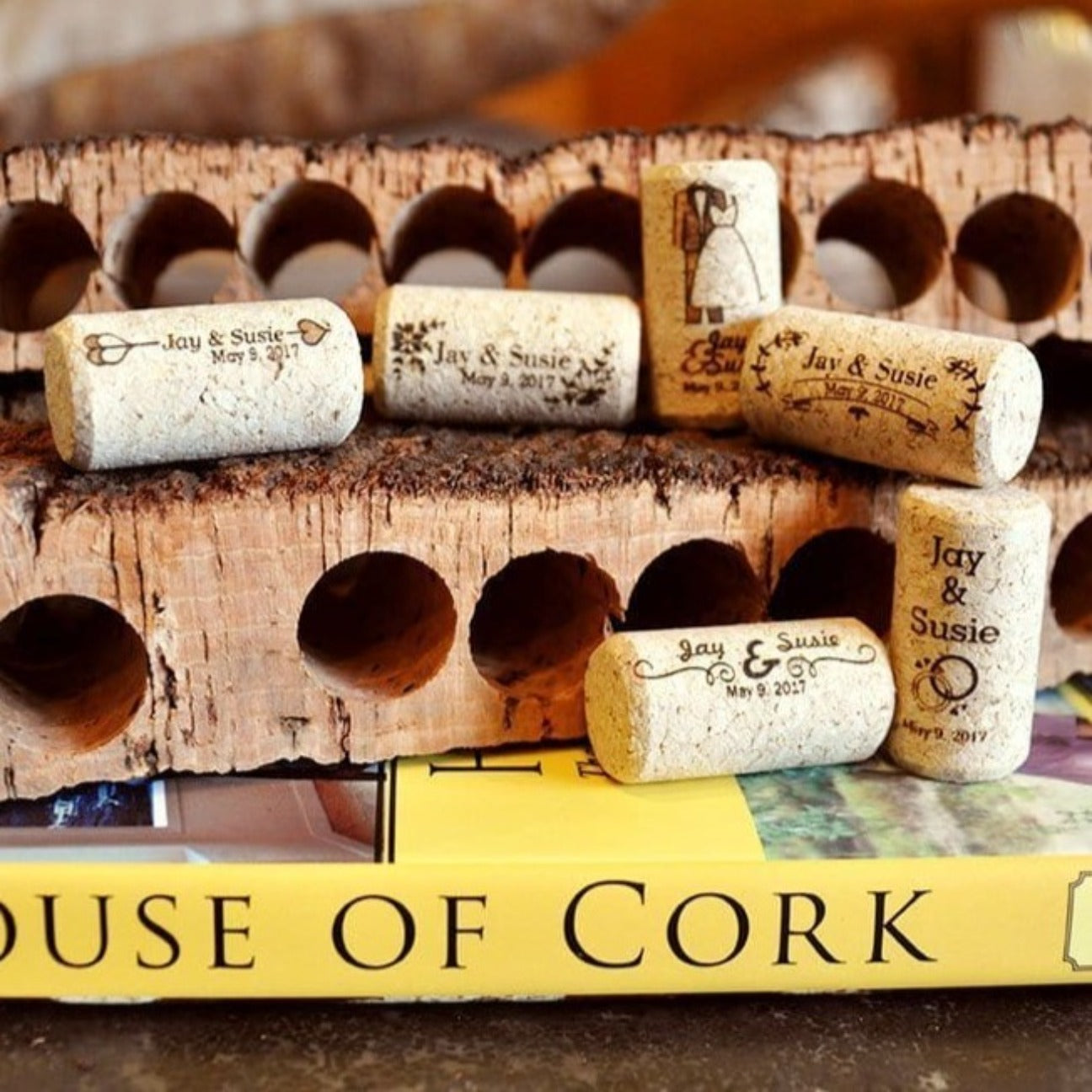 Six custom printed wine corks on a house of cork book with two cork bark punches in the background. Top left wine cork says "Jay & Susie May 7th 2017 with a cupid arrow. The wine cork second from the top left says "Jay and Susie May 7th 2017 with floral corner borders. The wine cork second from the right on the top row shows a man and woman wedding topper and says 